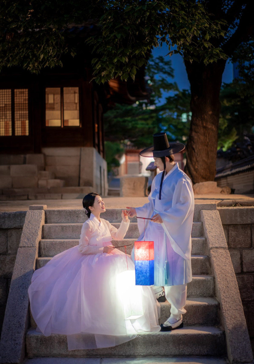 돌계단에 앉은 연분홍 한복의 여성과 곁에 선 남성이 손을 잡고 서로를 보며 웃는다. 청사초롱 불빛이 치마를 밝힌다