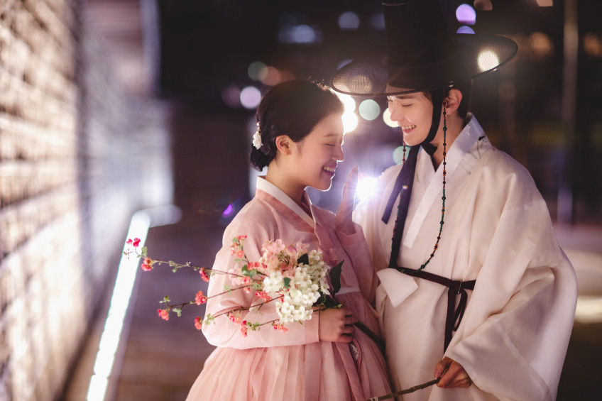 꽃가지를 든 연분홍 한복의 여성과 흰 한복의 남성이 밤길에서 이마를 맞대고 환하게 웃는다
