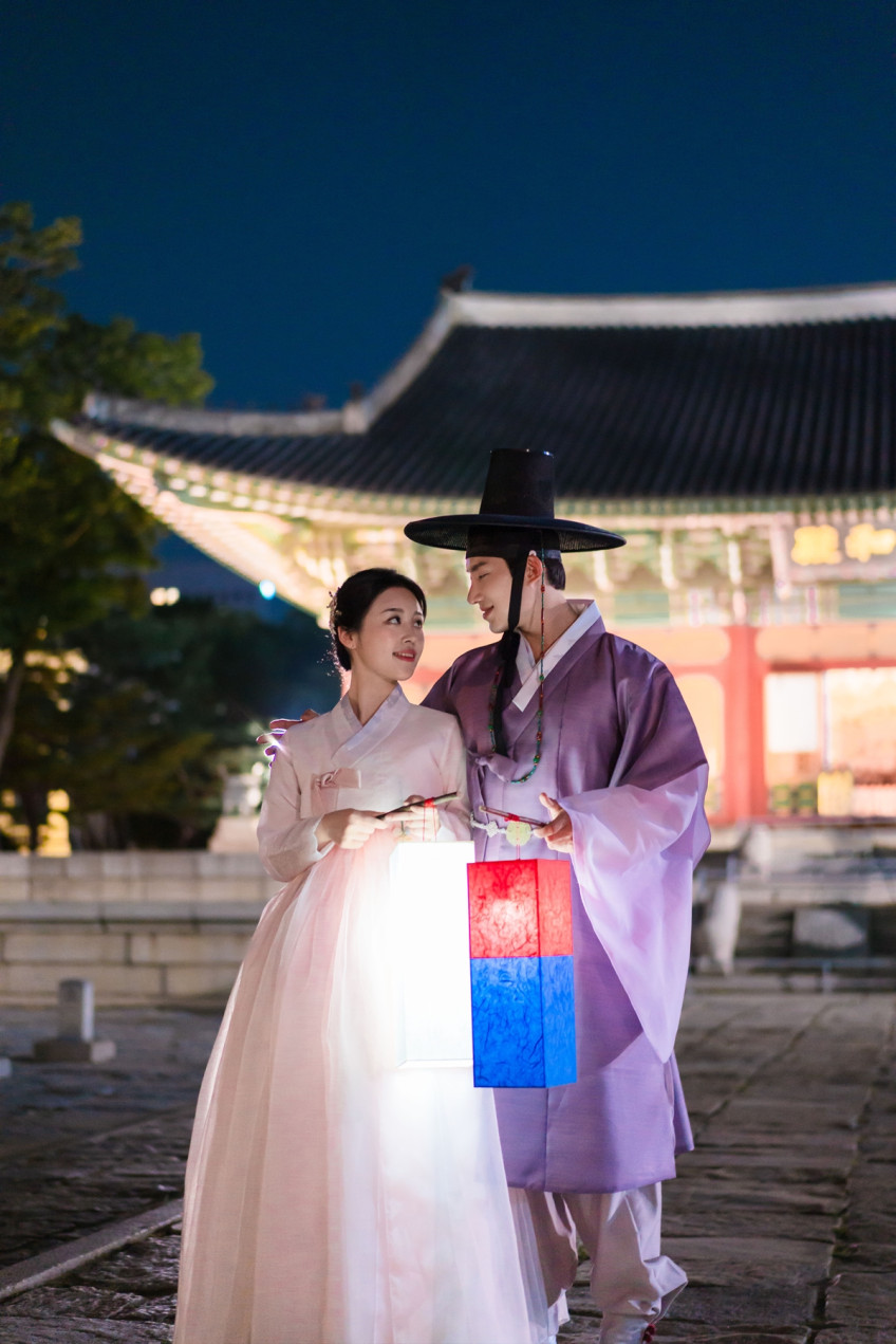 남청색 밤하늘 아래 기와 건물 앞에서 청사초롱을 든 연분홍 한복의 여성과 보라색 한복의 남성이 서로를 바라본다