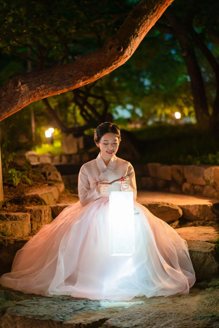 큰 나무 아래 돌계단에 앉아, 환하게 켜진 등을 두 손으로 들고 내려다보며 미소 짓는 연분홍 한복의 여성